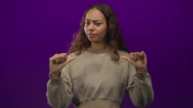 Stüdyoda iki başparmağı göğsünü gösteren, soluk eşofman üstü giyen ve kameraya bakarken kuşkulu ifadeler takınan genç bir kadın..