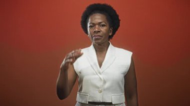 Kadın açık avuç içi bir jest sunuyor, gülümsüyor ve beyaz bir yelek giyerken kırmızı bir stüdyo arka planına karşı eline bakıyor; neşeli bir merak.