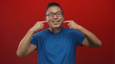 Erkekler kırmızı stüdyoda dişleriyle gülümserken, gözlük takarken ve mavi tişört giyerken yanaklarını işaret eder; neşeli neşe.