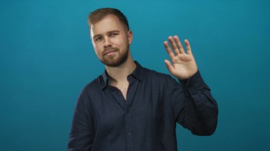 Deniz mavisi gömlekli ve sakallı bir adam görünür, açık yakalı ve kameraya bakan rahat duruş; dostça selamlaşma.