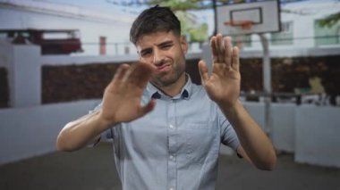 Açık mavi gömlekli genç İspanyol adam, açık hava sahasında güneş ışığı altında bir basketbol potasının yanında ellerini göğsünün üzerinden uzatıyor..