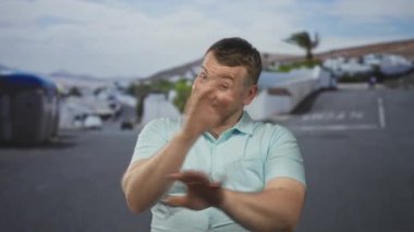 Açık mavi gömlekli bir adam beyaz binalar ve bir palmiye ağacı olan bir caddede avuçlarını ileri doğru sallamak ve bloklamak için el kaldırıyor..