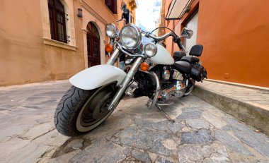 Beyaz kruvazör motosikleti dar bir kaldırım taşı caddesine park edilmiş, sıcak renkli binalar var.