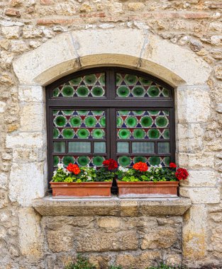 Yeşil camlı kemerli pencereler, tarihi bir taş duvarı kırmızı sardunyalarla süslüyor.