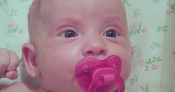 Een kleine schattige baby liggend in bed met een roze fopspeen in zijn mond.close-up — Stockvideo