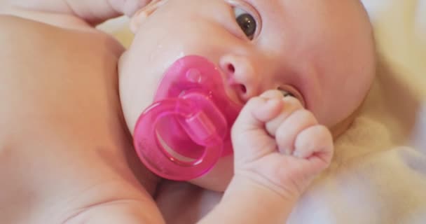 Porträtt av en söt bebis med en rosa napphållare i munnen.Ett litet barn med en förvånad blick ligger på lakanet.Närbild — Stockvideo