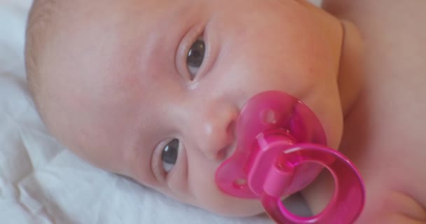 Porträtt av ett modernt barn. Söt baby spädbarn med en rosa napphållare i munnen ligger i sängen. närbild — Stockvideo