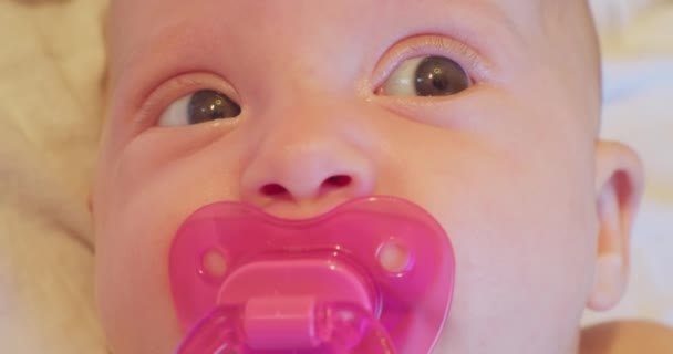 Gezicht van een kleine grappige en schattige baby met een fopspeen in zijn mond en een verrassende blik. close-up portret — Stockvideo