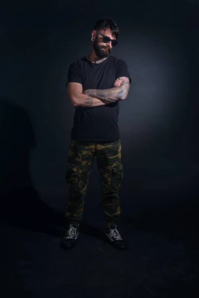 Bearded male model with tattoos and with crossed arms, dressed in a black t-shirt and sunglasses poses over black background.