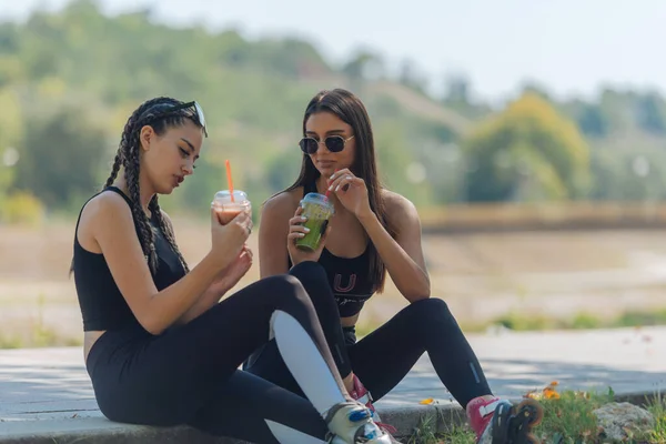 二人の女の子は スムージーとゴシップを飲んでローラースケートから休憩を取りながら公園に座っています 女の子の結合概念 — ストック写真