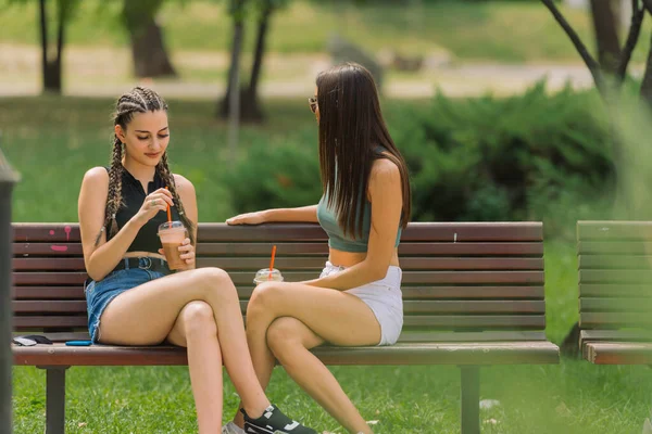 ベンチの屋外に座っている間に彼らのスムージーを飲んでいる2人の楽しい若い女性の公園の茂みを通して写真 — ストック写真