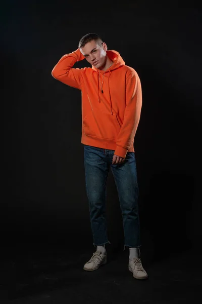 Young handsome man in jeans and orange hoodie posing while standing on a black background.