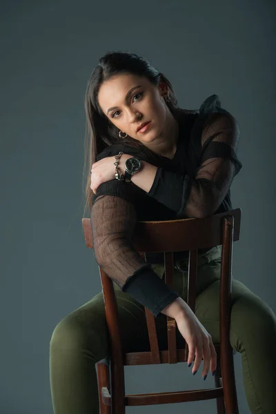 迷人的黑发模特坐在一张旧椅子上摆姿势 看着灰色背景的相机 — 图库照片