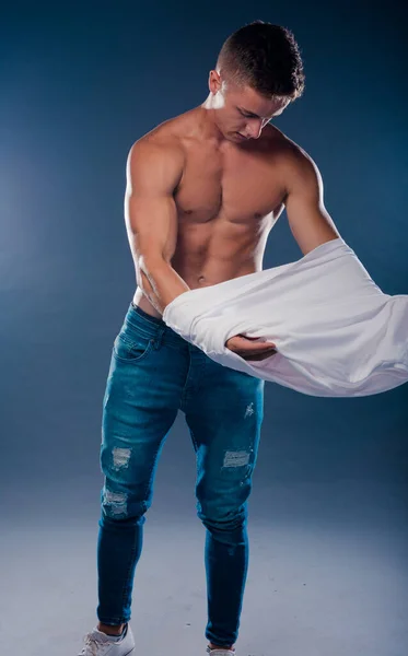 Handsome young muscular man shirtless wearing jeans, taking off white shirt on naked muscle torso