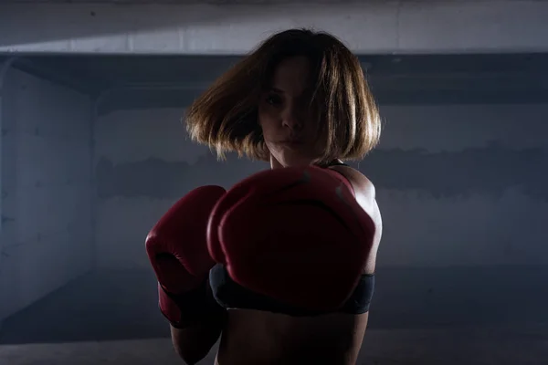 Silhouette portrait of young attractive fitness woman exercising punching with red boxing gloves in a garage