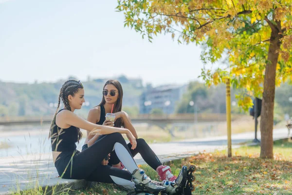 二人の女の子は スムージーとゴシップを飲んでローラースケートから休憩を取りながら公園に座っています 女の子の結合概念 — ストック写真