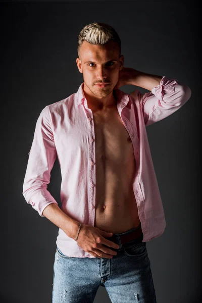 Cheerful pleased caucasian guy with blond hair dressed in elegant pink shirt smiling in good mood while posing in studio against dark background