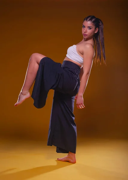 Full-body portrait of a young pretty girl posing in wide black pants and a white crop top with her leg up