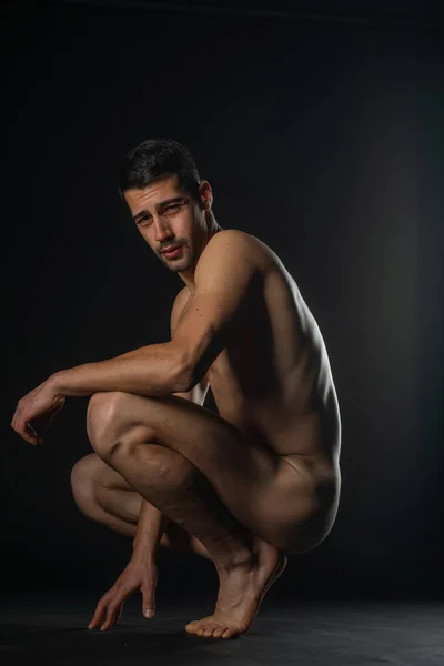 Portrait of a naked male model posing squatted on black studio background