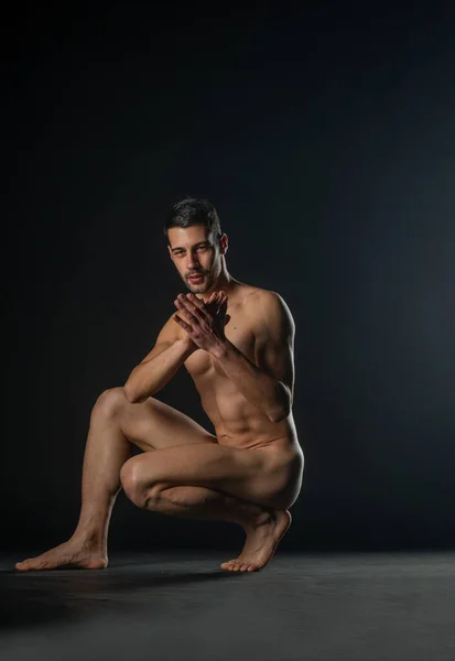 Portrait of a naked male model posing squatted on black studio background