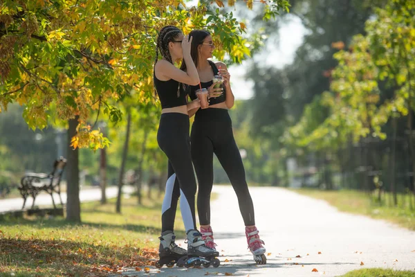 彼らのローラースケートに立っている間 公園で2人の魅力的な親友が話してスムージーを飲んでいます — ストック写真