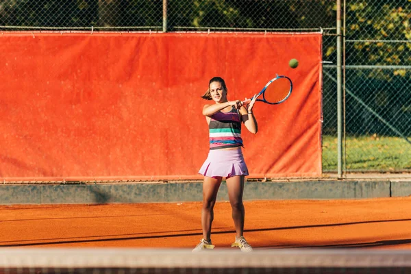 A young fit woman plays tennis outdoor on an orange tennis field early in the morning