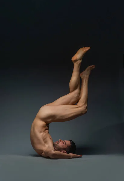 Upside down photo of a young naked man lying down on the floor in a dark studio with his legs up in the air