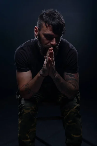 Closeup dark picture of a bearded male model in black t-shirt  sitting on a chair and praying with his tattooed hands