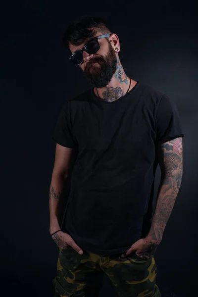 Bearded male model with tattoos and hands in his pocket, dressed in a black t-shirt and sunglasses poses over black background.