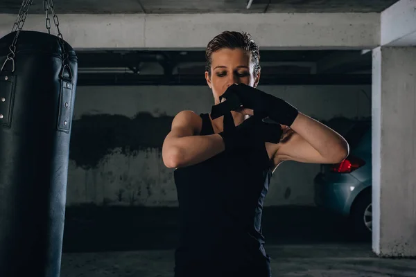 一个年轻强壮的女人用黑色绷带缠在手腕上准备在车库里打拳的画像 — 图库照片