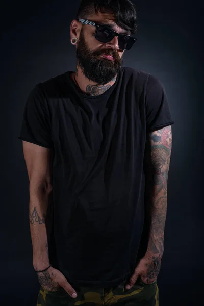 Bearded male model with tattoos and hands in his pocket, dressed in a black t-shirt and sunglasses poses over black background.