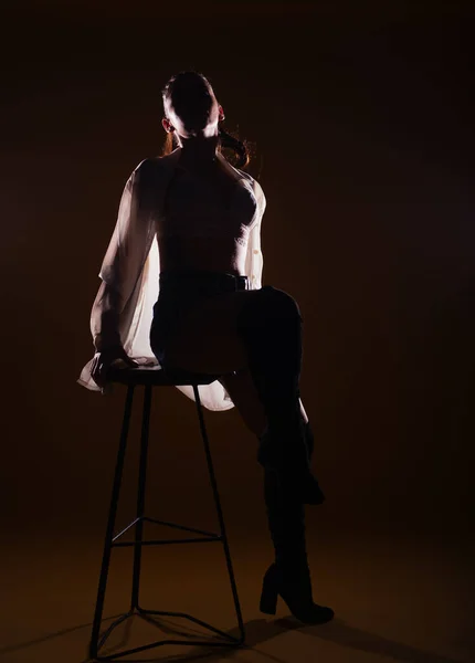 背の高い椅子を着てポーズをとるセンセーショナルな女性の暗い背景にフル長シルエットの肖像画 — ストック写真