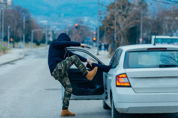 The car thief is pulling the car owner out of his car and trying to get the car while pointing a loaded gun at the drivers head.