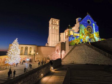 ASSISI, İtalya - 10 Aralık 2025: Assisi, İtalya 'da Aziz Francis Bazilikası' nın çarpıcı bir gece fotoğrafı, şenlikli Noel tahminleri ve yakınlardaki büyük, parlak bir Noel ağacı ile aydınlatılmış.