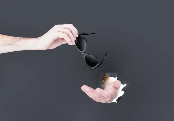 Male hand breaking through the black paper background and holding sun glasses — Stock Photo, Image