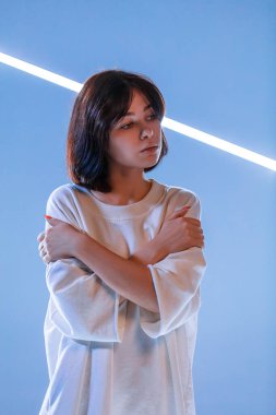 Kollarını vücudunun önünde kavuşturmuş genç bir kadın. Ciddi bir ifadesi var ve yumuşak mavi ışıkla çevrili, sakin ve yansıtıcı bir atmosfer yaratıyor..