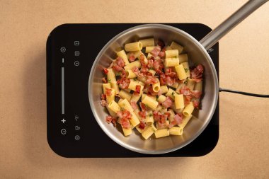 Makarna ve kıtır domuz pastırması, paslanmaz çelik bir tavada ince bir indüksiyon sobasında birlikte pişiriliyor. Dekor, temiz hatları olan çağdaş bir mutfak..