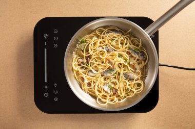 Spagetti, indüksiyon sobasında gümüş bir tencerede pişiriliyor. Sahne parlak bir mutfakta geçiyor, eriştelerin ve kullanılan taze malzemelerin altını çizerek..