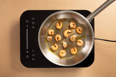 Sulu karidesler elektrikli ocakta gümüş bir tavada pişiriliyor. Etrafı nötr bir tezgahla çevrili sıcak bir mutfak ortamında..