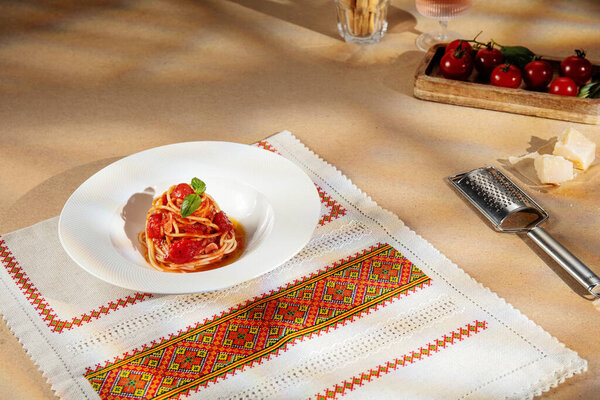 Plate of spaghetti with tomato sauce and basil on a traditional embroidered tablecloth under warm sunlight. Parmesan, tomatoes and wine create a cozy Mediterranean home-cooking mood.