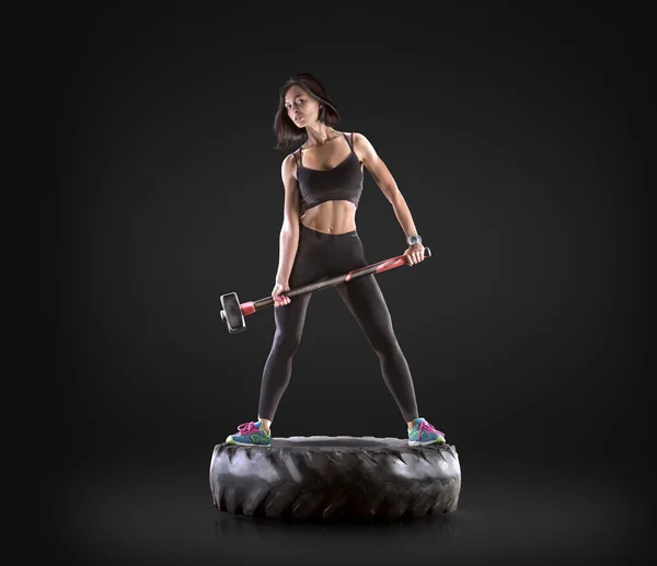 Атлетичная молодая женщина, занимающаяся фитнесом с молотком и шиной на фоне черной студии — стоковое фото