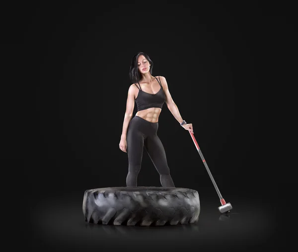 Атлетичная молодая женщина, занимающаяся фитнесом с молотком и шиной на фоне черной студии — стоковое фото