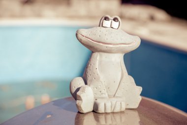 Small figurine of a cute frog at the edge of an empty swimming pool. Autumn concept clipart