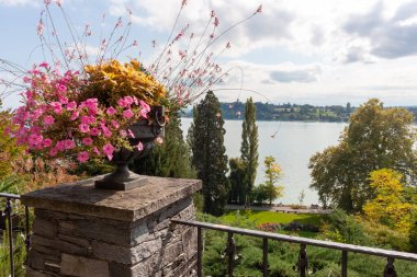Klasik taştan bir vazoda pembe petunyalar Mainau Adası 'nın teraslarından Constance Gölü' nün manzaralı manzarasını çerçeveliyor. Bulutlu bir gökyüzü altında yemyeşil ve uzak kıyılar var, Konstanz.