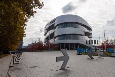 STUTTGART, GERMANY - 18 Ekim 2025: Stuttgart, Almanya 'daki Mercedes-Benz Müzesi' nin çarpıcı modern mimarisinin bulutlu bir gökyüzü altında yön işaretleri ve sonbahar ağaçlarıyla panoramik görünümü