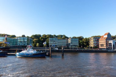 HAMBURG, Almanya - 12 AĞUSTOS 2024: Hamburg, Almanya 'daki Elbe Nehri' ne römorkör yanaştı. Modern rıhtım binaları ve mavi gökyüzünün altında manzaralı liman manzarası