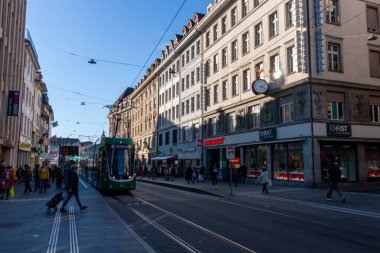 BASEL, SWitzERLAND - 16 ARALIK 2024: İsviçre 'nin Basel kentinde tarihi binalar ve açık bir gökyüzü altında perakende mağazalarıyla çevrili işlek bir ticari caddenin raylarından yeşil bir tramvay yaklaşıyor