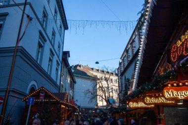 BASEL, SWitzERLAND - 16 Aralık 2024: Basel Noel Pazarı 'ndaki hareketli bir sokak sahnesi, tarihi binalar çerçeveli ve asılı ışıklar ve akşam gökyüzünün altında aydınlatılmış ahşap tezgahlarla süslenmiş