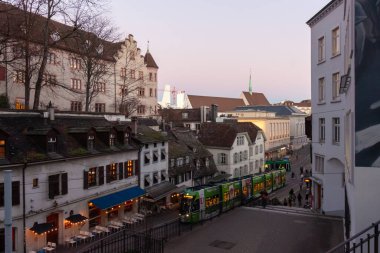 BASEL, SWitzERLAND - 16 Aralık 2024: Barfusserplatz yakınlarındaki eski şehir sokaklarında tarihi çatıları ve modern Roche Kuleleri 'nin arka planında alacakaranlıkta seyahat eden yeşil tramvay