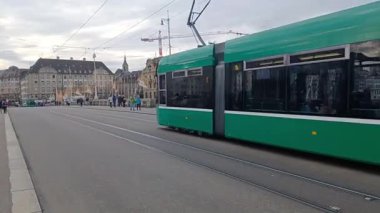 BASEL, SWitzERLAND - 10 ARALIK 2025: canlı yeşil bir tramvay Basel, İsviçre 'de kaldırımda yayalar ve Ren Nehri ile köprüden geçiyor.